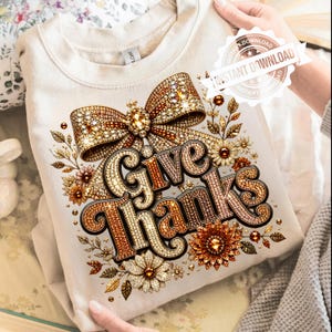 Può includere: Una felpa girocollo color crema con una stampa grafica di un grande fiocco dorato scintillante sopra il testo "Give Thanks" in un carattere decorativo. Il design include accenti floreali nelle tonalità dell'arancione, marrone e oro.