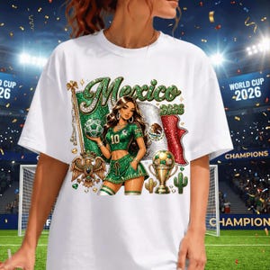 May include: A white t-shirt featuring a graphic of a person in a green soccer uniform holding a soccer ball. The design includes the text Mexico 2026, a Mexican flag, a golden trophy, a golden eagle emblem, and two small cacti on a stadium background.