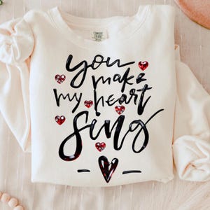 Può includere: Felpa color crema con la frase "You make my heart sing" in corsivo nero. Accenti di cuore rosso con strass sono sparsi intorno al testo e un cuore grafico è in basso.