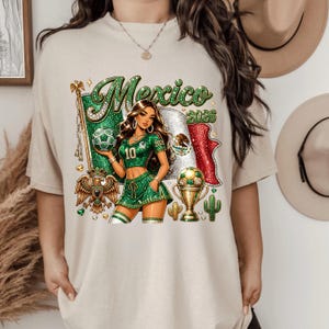 May include: A beige t-shirt featuring a graphic of a person in a green soccer uniform holding a soccer ball. The design includes the text Mexico 2026, a Mexican flag, a golden eagle emblem, a trophy, and cacti, all rendered in a glittery style.
