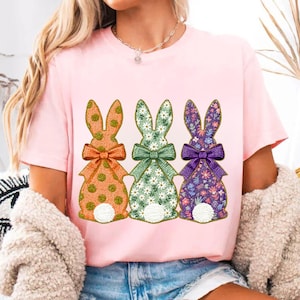 May include: Pink t-shirt featuring three Easter bunny designs. Each bunny has a different pattern: orange polka dots, white daisies on green, and purple floral. The bunnies have matching bows in coordinating colors.