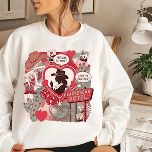 Puede incluir: Sudadera blanca con un diseño gráfico colorido. El diseño presenta una silueta en forma de corazón de una mujer con sombrero de vaquero, rodeada de botas de vaquero, naipes, cactus y las palabras "Heartbreak Hotel."
