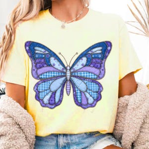 May include: A light yellow t-shirt featuring a large butterfly design. The butterfly has blue, purple, and white patchwork wings with a gingham pattern. The shirt is made of soft material.