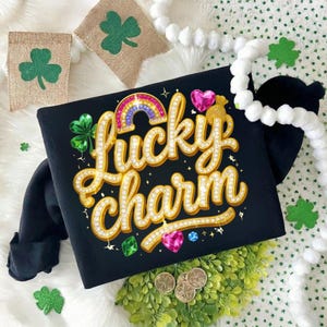 Puede incluir: Camiseta negra con la frase "Lucky Charm" en dorado, adornada con pedrería, un arcoíris y gemas en forma de corazón. El diseño incluye tréboles y monedas, evocando el tema del Día de San Patricio.