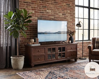 May include: A dark brown wooden media console featuring four drawers and two central glass-paneled cabinet doors with small round knobs. The rectangular unit sits on short feet and supports a flat-screen television displaying a lake landscape.