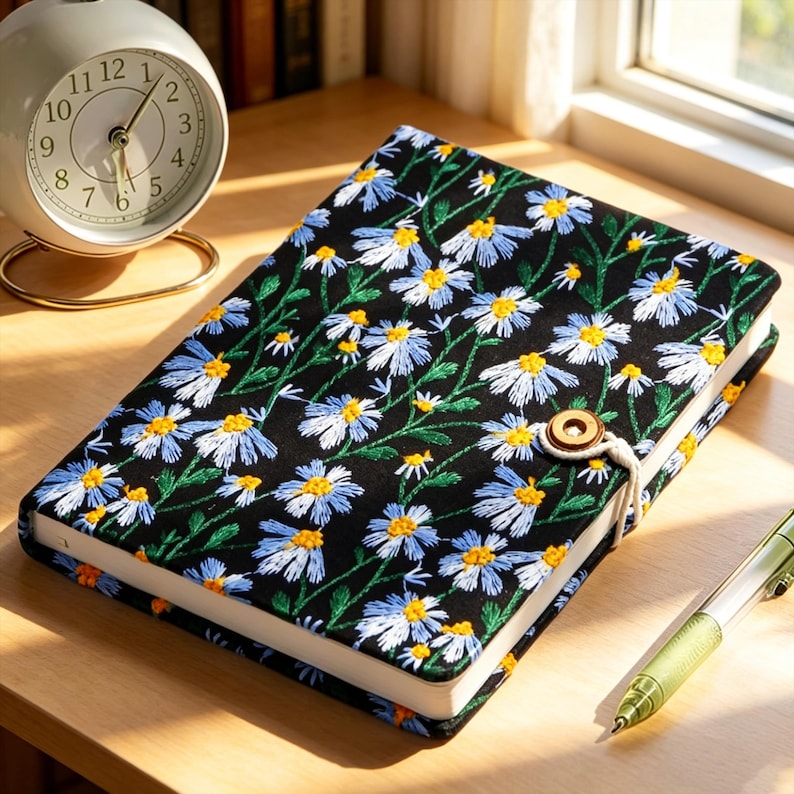 May include: A black journal with a floral daisy embroidery design. The cover features white and blue daisies with yellow centers and green stems. A wooden button and string closure is visible.