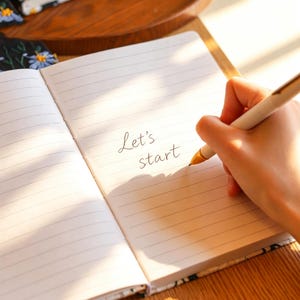May include: Open notebook with lined pages. The text "Let's start" is written on the page. A hand is holding a pen and writing on the page. The notebook has a floral design on the edges.