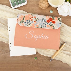 May include: A peach-coloured felt case embroidered with the name "Sophia" in white. The top flap has a floral pattern in red, blue, and green. A pen and a sheet of paper are next to the case.