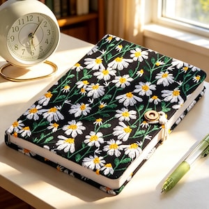 May include: A black journal with a white daisy floral embroidery design. The journal has a button closure and a cream-colored spine. A gold-colored alarm clock and a green pen are also on the table.