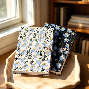May include: Two notebooks with floral embroidery. One notebook has a cream-colored cover with blue and yellow flowers, and the other has a black cover with blue and yellow flowers. Both notebooks have a wooden button closure.