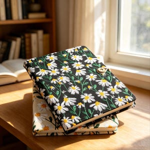May include: Two notebooks stacked on a wooden surface. The top notebook has a black cover with a white daisy pattern and a gold clasp. The bottom notebook has a beige cover with a daisy pattern. The notebooks are near a window and a bookshelf.