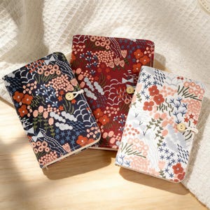 Könnte beinhalten: Drei floral bestickte Notizbuchhüllen in Marineblau, Burgunderrot und Weiß. Jede Hülle hat ein einzigartiges Muster aus Blumen und Blättern in Orange-, Rosa- und Grüntönen. Sie werden mit einem Holzknopf und einer Schlaufe verschlossen.