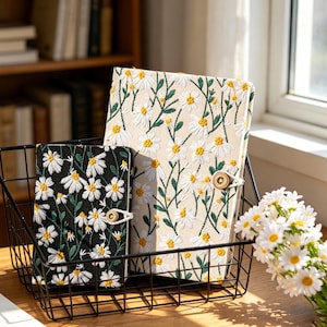 May include: Two daisy-embroidered journals in a black wire basket. One journal has a black background, the other is cream-colored. Both have white daisies with yellow centers and a wooden button closure. A bouquet of daisies sits nearby.