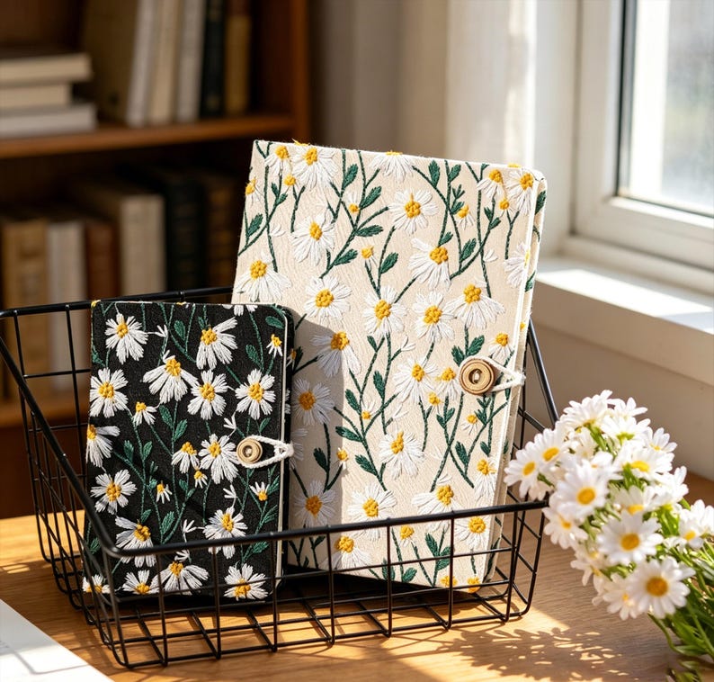 May include: Two daisy-embroidered journals in a black wire basket. One journal has a black background, the other is cream-colored. Both have white daisies with yellow centers and a wooden button closure. A bouquet of daisies sits nearby.