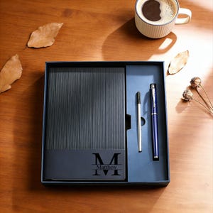 May include: A dark blue gift set containing a notebook with vertical lines, two pens, and a box. The notebook is engraved with the name "Matthew" and the initial "M". A cup of coffee is placed on a wooden surface nearby.