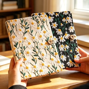 May include: Two embroidered journals with daisy patterns. One journal has a cream-colored background, and the other is black. Both have white daisies with yellow centers and green stems. Each journal has a wooden button closure.