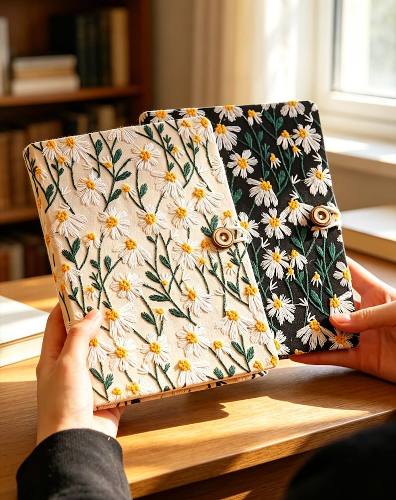 May include: Two embroidered journals with daisy patterns. One journal has a cream-colored background, and the other is black. Both have white daisies with yellow centers and green stems. Each journal has a wooden button closure.