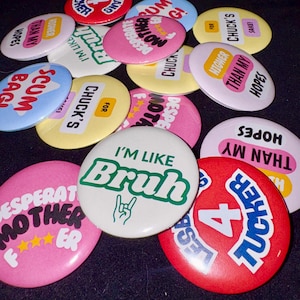 Op de afbeelding: Een verzameling ronde buttons in verschillende kleuren, waaronder rood, roze, geel, wit en blauw. Elke button is voorzien van vetgedrukte tekst met zinnen zoals "I'm Like Bruh", "Desperate Mother F***er", "Higher Than My Hopes", "For Chuck's Sake" en "Lesbians 4 Tucker".