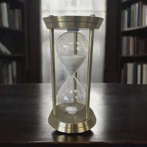 May include: An hourglass with a brass-colored metal frame and clear glass bulbs. The top bulb is filled with white sand, and the sand is flowing into the bottom bulb. The hourglass is sitting on a dark wooden surface.