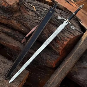 May include: A long sword with a textured, silver-toned blade, a black grip, and a metallic crossguard. The sword features a decorative pommel and is accompanied by a black leather scabbard with visible stitching and a thin, adjustable brown strap.