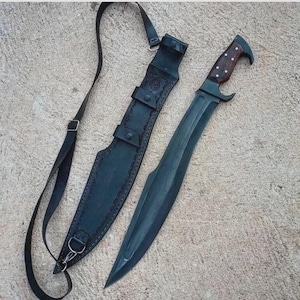 Può includere: Un coltello grande con lama scura, manico in legno e fodero in pelle nera. Il coltello ha una lama curva e un manico decorativo con piccoli punti bianchi. Il fodero ha una tracolla e passanti per cintura.