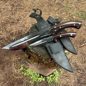 May include: Two large hunting knives with polished steel blades and dark wood handles with white accents. The knives are displayed with black leather sheaths and a leather belt attachment, set on a tree stump.