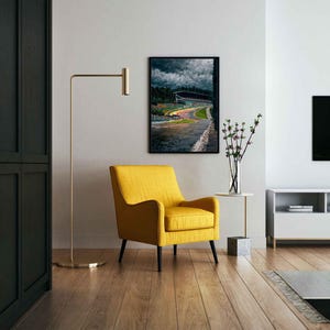May include: A contemporary living room with a framed race track print, a yellow armchair with black legs, and a gold floor lamp. The room has wooden flooring and a white side table with a vase of flowers.