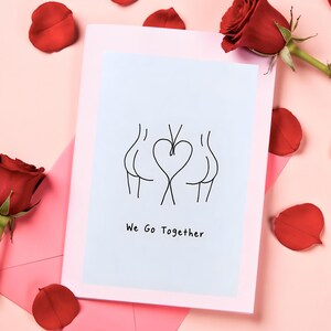May include: A greeting card with a light blue background and the text "We Go Together". The card features a black line drawing of three figures with a heart shape in the middle. Red rose petals and roses are scattered around the card.