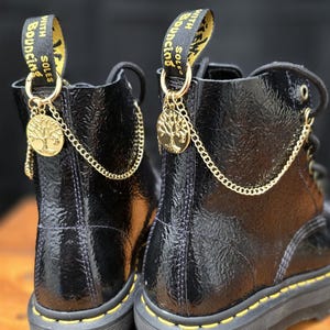 Puede incluir: Un par de botas negras de charol con suela de plataforma gruesa. Cada bota presenta un dije dorado con un diseño de árbol, suspendido por una cadena. Las trabillas de las botas tienen las palabras "WITH BOUNCING SOLES".