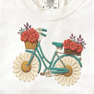 Könnte beinhalten: Cremefarbenes T-Shirt mit einem gestickten Design eines türkisfarbenen Fahrrads mit Gänseblümchenrädern und Körben mit roten Blumen. Das Shirt hat einen Rundhalsausschnitt und kurze Ärmel. Das Etikett lautet "COMFORT COLORS".