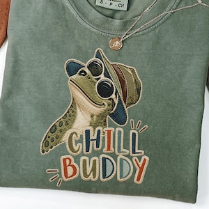 May include: An olive green t-shirt featuring an embroidered frog wearing sunglasses and a hat, with the text "CHILL BUDDY" below. A gold necklace with a pendant is draped across the shirt. The shirt is a small size.