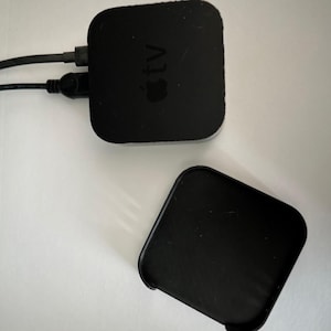 May include: A black Apple TV device with a connected power cord and a separate black remote. The Apple TV has the Apple logo and the word "tv" printed on it. The devices are on a white surface.