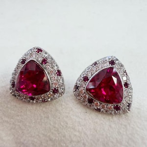 May include: A pair of triangular stud earrings featuring a large, faceted deep red centre stone surrounded by a halo of smaller, clear white stones and interspersed with tiny, round, deep red accent stones set in a bright silver-toned metal.