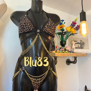 Op de afbeelding: Een zwarte mannequin toont een tweedelige lingerieset. De set heeft een top en broekje met luipaardprint, verbonden door zwarte bandjes en gouden kettingen. De tekst "Blu83" is zichtbaar op het kledingstuk.