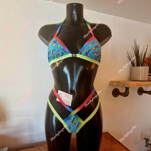 May include: A two-piece swimsuit displayed on a black mannequin. The halter top and bottoms feature a blue background with a colorful pattern. The edges are trimmed with rainbow and neon green accents. A small gem is at the center of the top.