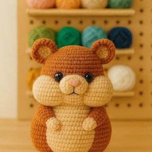 Puede incluir: Un juguete de hámster hecho a mano con ganchillo en tonos marrón y crema, con ojos negros y una nariz rosa. El peluche tiene mejillas grandes y pequeñas patas. En el fondo se ven bolas de hilo de colores, lo que sugiere un tema artesanal.