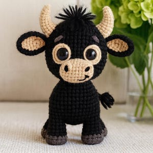 Peut inclure: Un taureau noir fait à la main au crochet avec des accents beiges. Le taureau a de grands yeux expressifs, de petites cornes et une touffe de cheveux touffue. Le taureau est assis sur une surface neutre, avec des fleurs vertes en arrière-plan. Une peluche mignonne et fantaisiste.