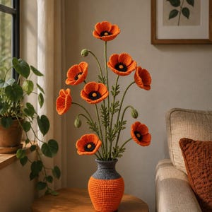 Pode incluir: Um buquê de papoulas laranja de crochê feitas à mão com centros pretos e caules verdes em um vaso bicolor laranja e cinza. O vaso está em uma mesa de madeira, com uma janela e uma impressão botânica emoldurada no fundo.
