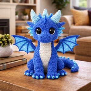 Może przedstawiać: Ręcznie robiony niebieski smok z szydełka z dużymi czarnymi oczami, jasnoniebieskimi skrzydłami i rogami. Smok ma biały brzuch i siedzi na drewnianym stole. Fantazyjny przedmiot o tematyce fantasy.