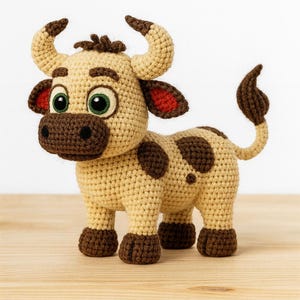 Op de afbeelding: Een gehaakte koeienfiguur in beige, bruin en rood. De koe heeft grote groene ogen, bruine vlekken en kleine hoorns. De staart is gekruld. Het handgemaakte speelgoed staat op een houten oppervlak.