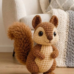 Puede incluir: Un juguete de crochet de ardilla hecho a mano en tonos marrón y beige. El peluche tiene una cola esponjosa, orejas pequeñas y ojos negros grandes. Está de pie, con los brazos a los lados. El fondo incluye una silla color crema y una manta decorativa.