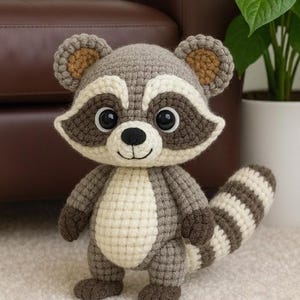 Könnte beinhalten: Gehäkeltes Waschbär-Plüschtier. Der Waschbär hat einen grau-weißen Körper mit gestreiftem Schwanz, dunkelbraunen Akzenten und großen, ausdrucksstarken Augen. Es steht auf einer hellen Oberfläche. Ungefähr 25 cm groß.