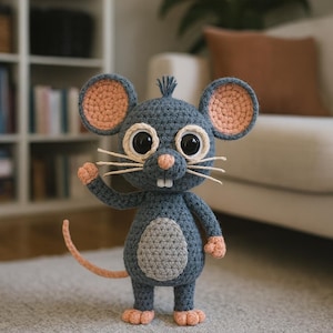 Könnte beinhalten: Handgefertigte Häkelmaus in Grau-, Pfirsich- und Cremetönen. Die Maus hat große, ausdrucksstarke Augen, einen langen Schwanz und eine freundliche Pose. Das Spielzeug steht auf einem grauen Teppich.