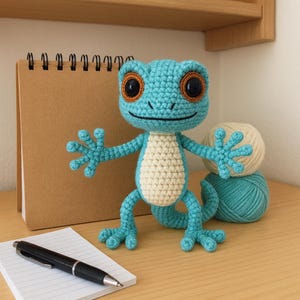 Peut inclure: Un gecko crocheté à la main, turquoise et crème, avec de grands yeux bruns. Le gecko a les bras tendus. Un bloc-notes, un stylo et des pelotes de laine sont en arrière-plan.