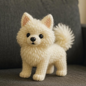May include: A handmade, cream-colored crochet dog figurine with a fluffy texture. The dog has black eyes, a black nose, and pink accents on its ears. It is standing on a gray surface, showcasing a detailed, artisanal craft.