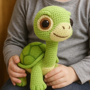 May include: A handmade crochet turtle toy in shades of green and cream. The turtle has large brown eyes, a smiling face, and a detailed shell. The toy is soft and plush, perfect for a child's playtime or nursery decor.