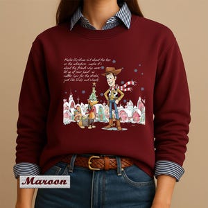 May include: A maroon long-sleeve sweatshirt featuring a graphic of Woody and Slinky Dog from Toy Story in a snowy village setting. The text reads: Maybe Christmas isn't about the toys or the adventure, maybe it's about the friends who never let go of your hand.