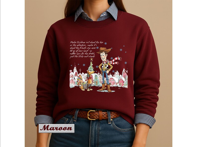 May include: A maroon long-sleeve sweatshirt featuring a graphic of Woody and Slinky Dog from Toy Story in a snowy village setting. The text reads: Maybe Christmas isn't about the toys or the adventure, maybe it's about the friends who never let go of your hand.