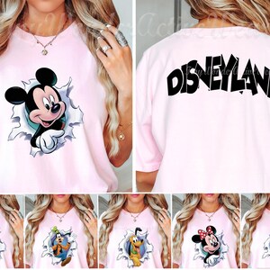 May include: A light pink t-shirt featuring a graphic of Mickey Mouse emerging from a torn paper effect on the front and the word Disneyland printed in large black letters across the upper back. The design includes various Disney character options.