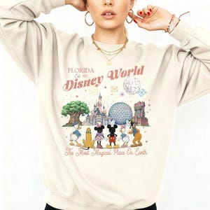 May include: A cream-colored crewneck sweatshirt featuring a graphic print of Disney World landmarks and characters. The text reads Florida Est 1971 Disney World The Most Magical Place On Earth. The design includes Mickey, Minnie, Donald, Daisy, and Pluto.